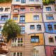 Šareno pročelje Hundertwasserove kuće u Beču s valovitim plohama u ružičastoj, plavoj i žutoj boji, prozorima različitih oblika i zelenilom na pročelju