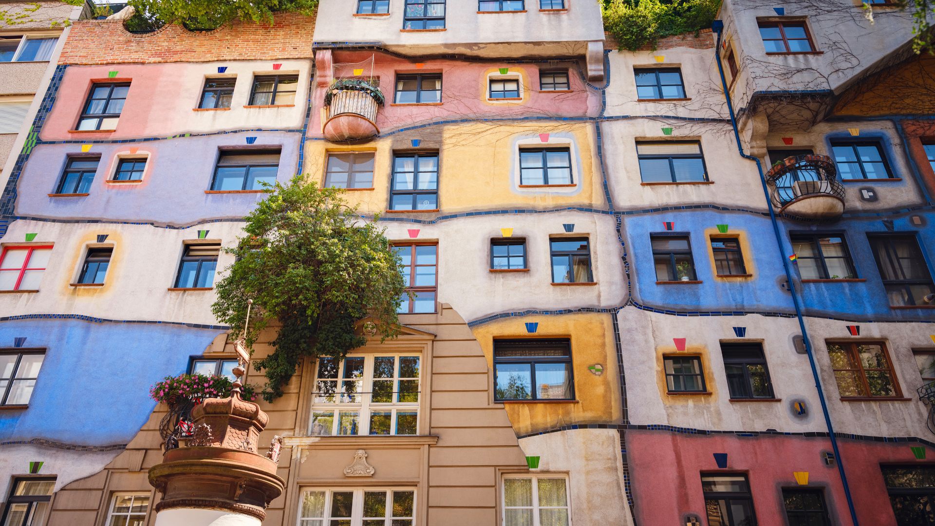 Šareno pročelje Hundertwasserove kuće u Beču s valovitim plohama u ružičastoj, plavoj i žutoj boji, prozorima različitih oblika i zelenilom na pročelju